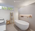 Modern bathroom with a freestanding bathtub, double sink vanity, wooden shelf, and decorative items on a built-in shelf. Natural light from large windows.