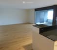 Empty room with light wooden flooring, white walls, and a black countertop in the foreground. A sliding glass door leads to a balcony area with brick walls and a view of the outside.