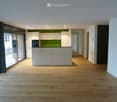 Empty room with light wooden flooring, white walls, and a small kitchen area with white cabinets and a green backsplash visible in the background.