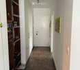 A narrow hallway with wooden tiled flooring, white walls, and a built-in shelving unit on the left side. A closed door is visible at the end of the hallway.