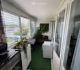 Balcony with green carpet, wooden floor, large windows with blinds, sofa, table, chair, refrigerator, microwave, printer, and potted plants.