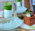 A small, elegantly presented dessert on a plate, accompanied by a glass of water and a small drink in a clear glass. The dessert features a brown base, green topping, and a small dollop of cream with berries.