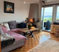 Modern living room with wooden flooring, grey sofa, coffee table, fireplace, and scenic view through large windows.