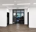 Empty room with wooden flooring, white walls, and recessed lighting. A glass door leads to an adjacent office space with desks, chairs, and office equipment.