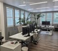 Modern office space with white desks, chairs, multiple computer screens, laptops, and potted plants near large windows with blinds.