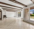 Bright, open space with exposed wooden beams, tiled floor, and large glass doors leading to an outdoor area. Features a built-in brick fireplace and white walls.