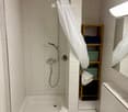 Modern bathroom with white tiled walls and floor, featuring a bathtub, toilet, sink, mirror, and a wooden shelf holding towels and toiletries.