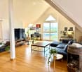 Bright living room with wooden flooring, large windows, a sectional sofa, coffee table, TV stand, piano, and decorative plants.