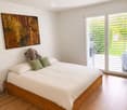 Modern bedroom with a wooden bed, white bedding, green pillows, an elephant plush toy, and a large abstract artwork on the wall. Large windows with blinds offer a view of the garden.