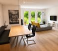 Modern living room with a wooden table, black leather bench, grey sofa with yellow cushions, black chairs, floor lamp, and a TV on the wall. Large windows provide a view of a green garden.