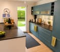 Modern kitchen with teal cabinets, dark countertops, and a large island. Features a sink, coffee maker, cutting board, bowl of apples, clock, and books on a shelf.