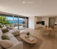 Modern open-plan living and dining area with light wood flooring, large sliding glass doors leading to a balcony with sea view, a beige sectional sofa, a round coffee table, a dining table with chairs, and decorative plants.