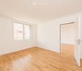 Empty room with light wooden flooring, white walls, and a large window showing a view of neighboring buildings. An open door leads to another space.