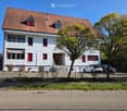 White painted house, brown roof, red shutters, cars parked in front, trees, paved driveway