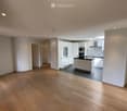 Open-plan space with light wooden flooring, white walls, and a modern kitchen area featuring white cabinets, a black countertop, and built-in appliances.