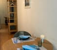 A small, neatly arranged dining area with a round wooden table set for two, featuring plates, wine glasses, cutlery, and a lit candle. A wooden shelving unit is visible in the background.