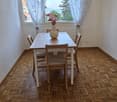 Dining room with wooden herringbone floor, white dining table, four chairs, and a vase with flowers on the table. Large window with white curtains and a view of greenery outside.