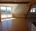 Bright, empty attic room with wooden floors, large windows, and exposed wooden beams. Features a balcony area with outdoor view.