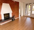 Empty room with wooden flooring, a brick fireplace with a white mantel, and large windows with sliding glass doors leading to an outdoor area.