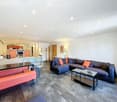 Modern open-plan living and dining area with a grey sectional sofa, orange throw pillows, a black coffee table, a pink bench, and a small kitchen area with orange cabinets and stainless steel appliances.