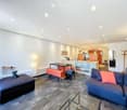 Modern living room with a grey sectional sofa, glass coffee table, colorful bench seating, and a small table with stools. The space features a sleek kitchen area with orange cabinets and a white countertop.
