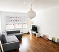 Modern living room with wooden flooring, large windows, grey sectional sofa, coffee table, TV, and decorative artwork.
