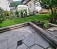 Garden area with stone-paved patio, wooden log border, small garden bed with plants, and a wooden fence surrounding the space.
