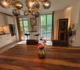 Open-plan kitchen and dining area with modern white cabinets, a wooden dining table, and a vase of flowers in the center. The space features large windows with curtains, a chandelier, and a small seating area with stools.