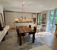 Modern open-plan living and dining area with a wooden dining table, chairs, kitchen cabinets, and a sleek kitchen sink. The space features a large mirror, decorative vases, and a potted plant, with natural light streaming in from large windows and sliding