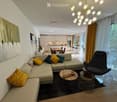 Modern living room with a sectional sofa, colorful throw pillows, a coffee table, a round side table, a modern chair, and a dining area with a table and chairs in the background.