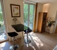 Bright room with wooden flooring, large windows, two black stools, a glass table, a lamp, a wooden cabinet, a mirror, and a potted plant.