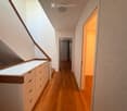 Modern hallway with wooden flooring, white walls, built-in storage unit with drawers, and a skylight providing natural light. The hallway leads to another room with a visible door and window.