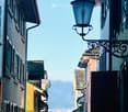 A narrow street lined with colorful buildings, featuring ornate street lamps and a view of distant rooftops and mountains.
