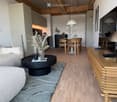 Modern living room with a grey sofa, coffee table, dining area, and kitchen in the background. Features wooden flooring, pendant lights, and a sleek TV stand.