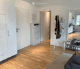Bright living room with wooden flooring, a grey sofa, a large rug, a white wardrobe, a shoe rack, and a visible kitchen area with cabinets and appliances.