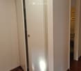 A narrow hallway with wooden flooring, white walls, and a closed white door on the left leading to another room. A partially open door on the right reveals a glimpse of a laundry area with a washing machine.