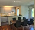 Modern dining area with a wooden dining table and six black chairs, adjacent to a sleek kitchen with white cabinets and stainless steel appliances.