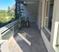 Balcony with tiled floor, metal railing, and a view of greenery. Cleaning equipment is placed near the railing.