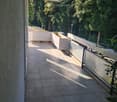 Balcony with tiled floor, modern railing, and a covered section, overlooking a green area with trees and parked cars.