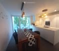 Modern kitchen with white cabinets, a central island, and a dining area with a wooden table and chairs. The space features pendant lighting and large windows overlooking an outdoor area.