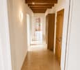 Clean, modern hallway with beige tiled flooring, white walls, and wooden doors. A staircase with wooden railings is visible in the background, along with a wall-mounted light fixture on the left side.