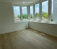 Empty room with light wooden flooring and large windows offering a view of rooftops and trees.