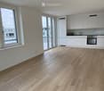 Empty kitchen with white cabinets, wooden flooring, large windows, and a small sink area.