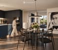 Modern dining area with a round glass table, four chairs, vase with flowers, books, candle, and cup. Adjacent living area features a sofa, coffee table, and plant. Kitchen area visible with dark cabinets, coffee machine, oven, and fridge.