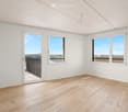 Empty room with white walls, wooden floor, and large windows offering a scenic view. Features a sliding door leading to a balcony.