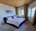 Modern bedroom with a large bed, nightstand, lamp, fan, and a scenic view through large windows with curtains.