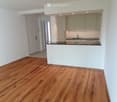 Empty room with wooden flooring, white walls, and a white kitchen area with cabinets and a countertop.