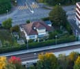 Aerial view of a residential area with a two-story house, a multi-story apartment building, a railway track, and surrounding greenery.