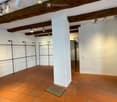 Empty room with exposed wooden beams, terracotta tiles, and white walls. Features a central pillar and several hanging spotlights.