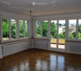 Bright, unfurnished room with large windows and a balcony. The room features wooden flooring, white walls, and radiators along the walls.